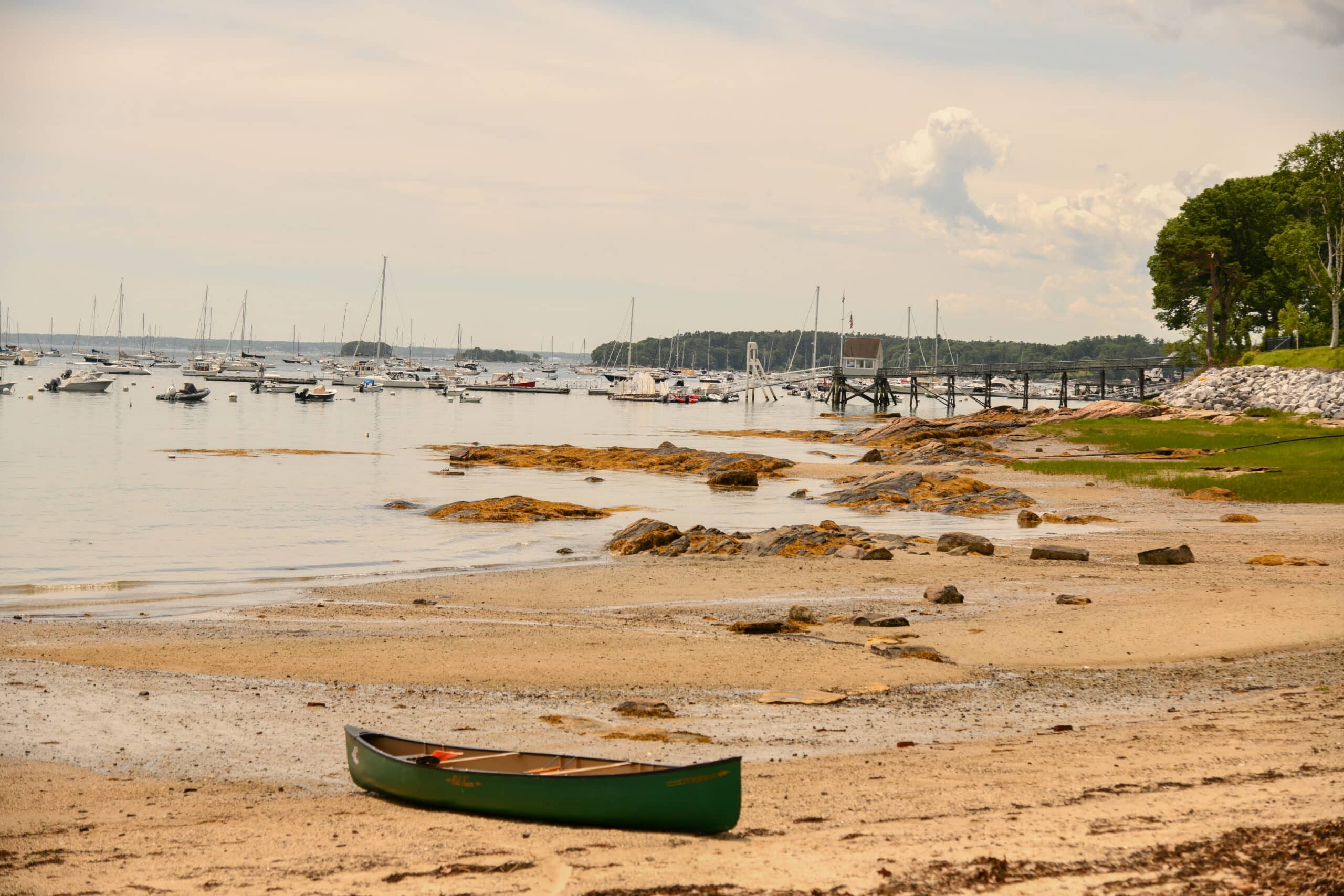 Rocky beach with a green canoe, sailboats moored in a harbor, and a small dock