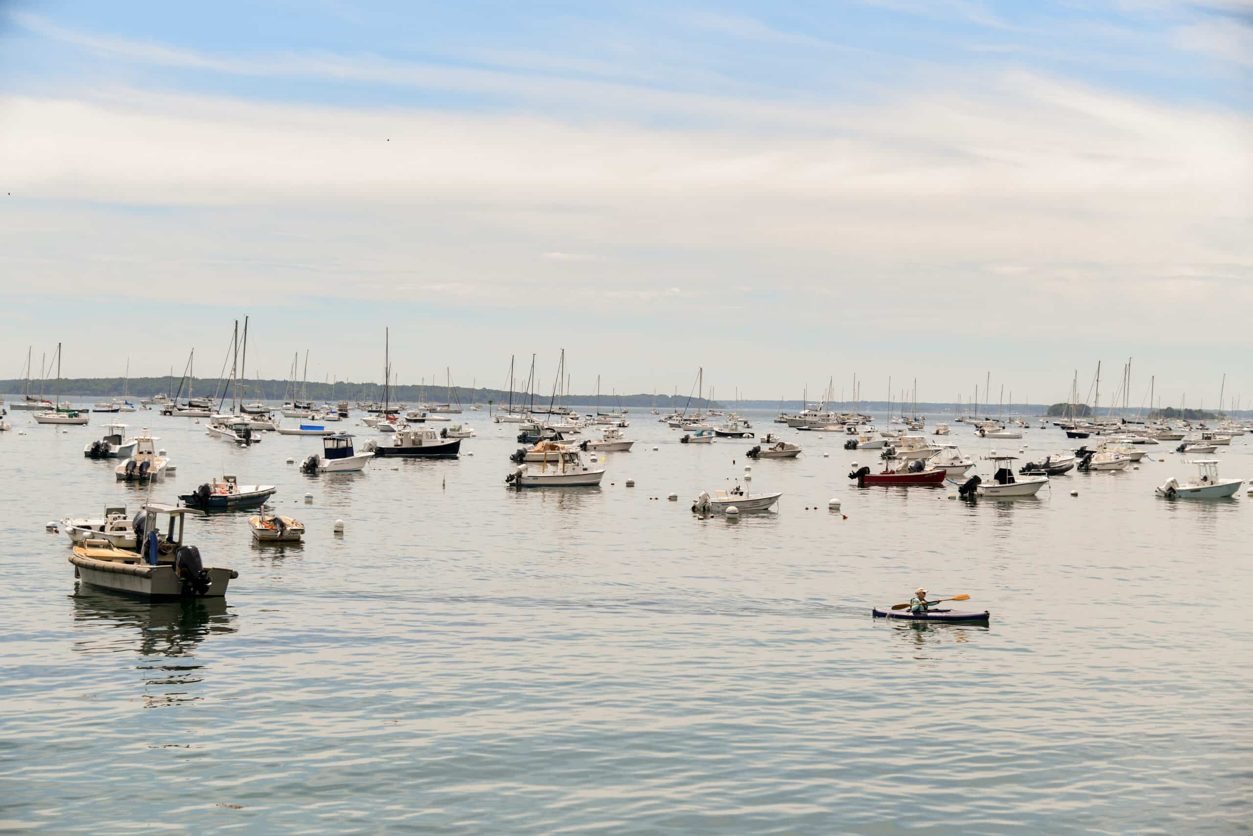 Harbor filled with sailboats and motorboats, with a kayaker paddling across calm water
