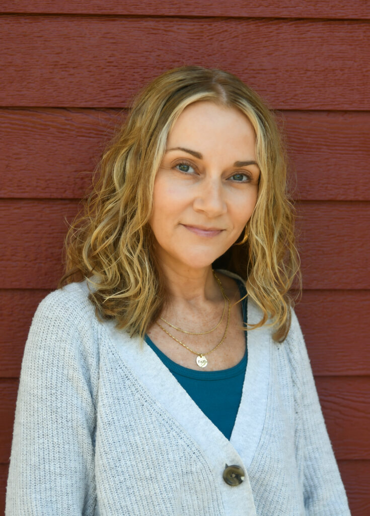 Woman with wavy blonde hair wearing a light cardigan and necklace, posing in front of a red wooden wall