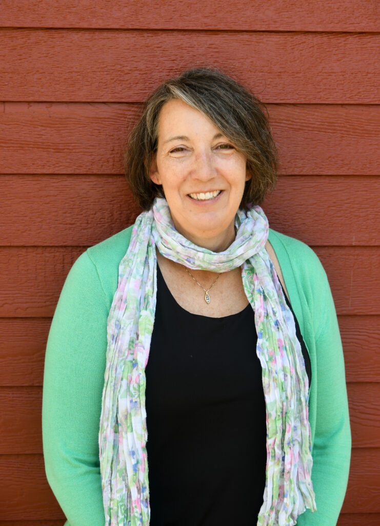 Smiling woman wearing a green cardigan and floral scarf in front of a red wooden wall