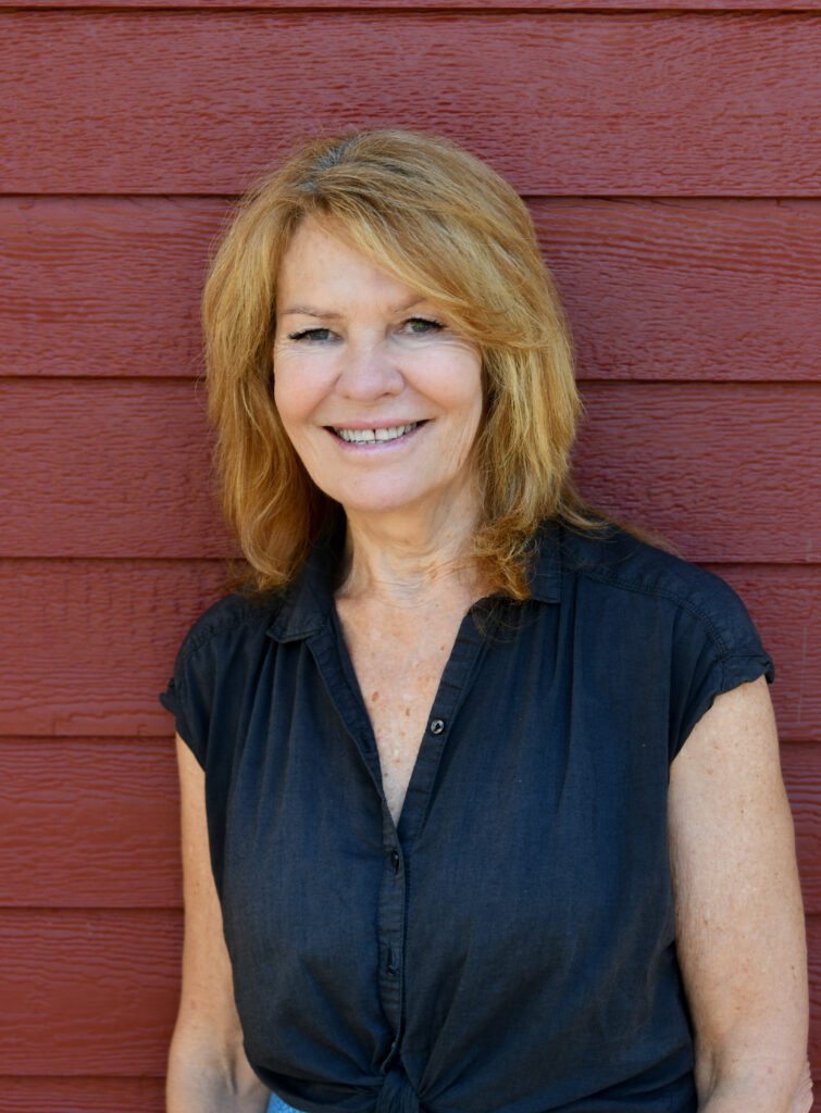 Smiling blonde woman in a black blouse standing in front of a red wooden wall
