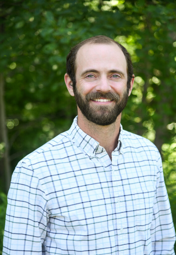 Smiling bearded man wearing a white plaid shirt outdoors against a leafy green background