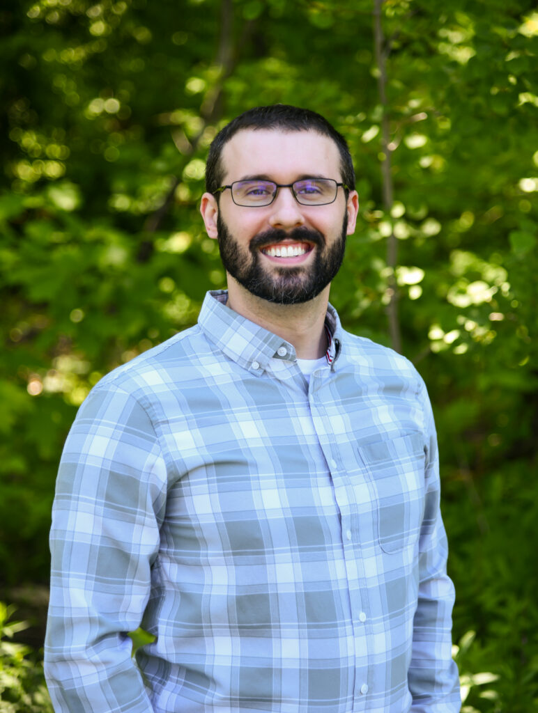 Smiling bearded man wearing glasses and a light plaid shirt outdoors against a leafy green background