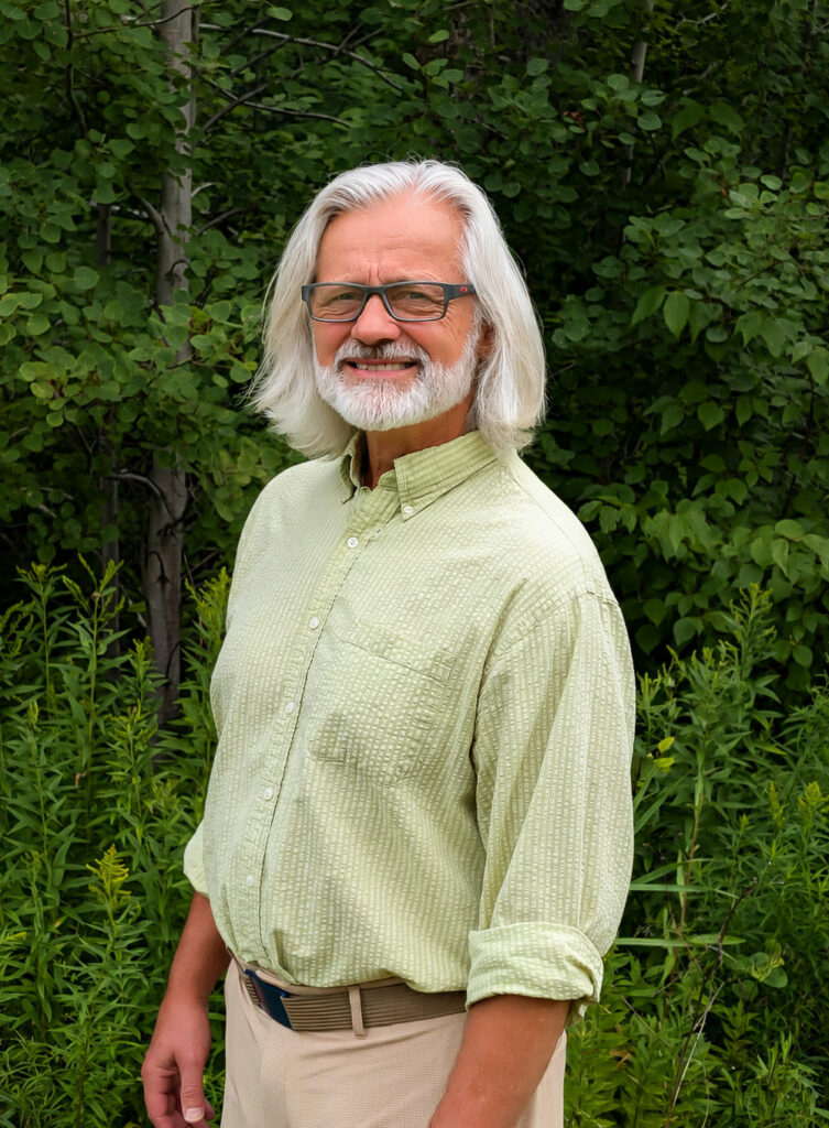 Smiling older man with long white hair and glasses standing in a leafy garden