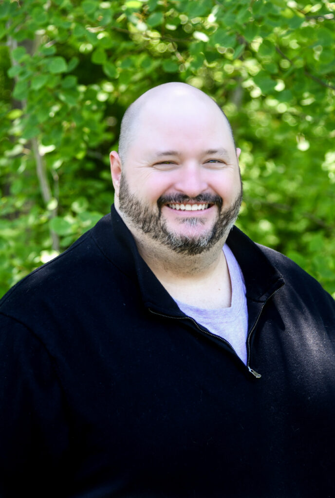 Smiling bald man with a beard wearing a black jacket outdoors against leafy greenery