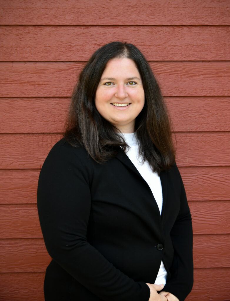 Smiling woman with long dark hair wearing a black blazer in front of a red wooden wall