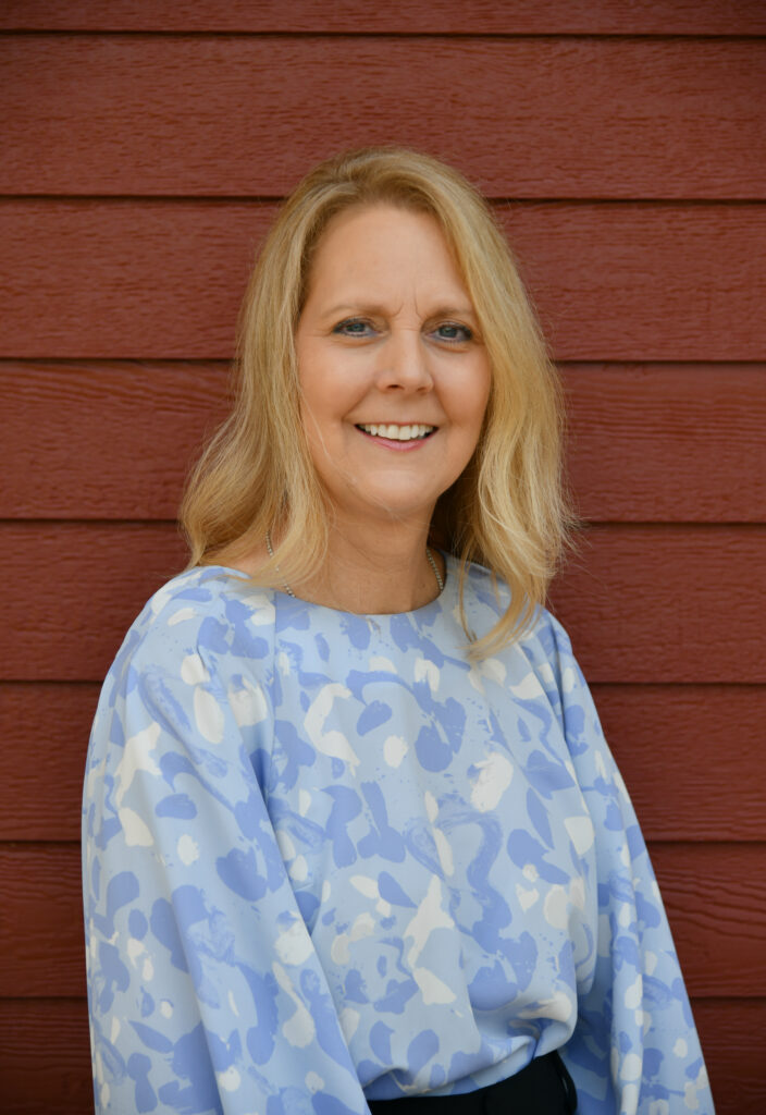 Smiling woman with long blonde hair wearing a light blue patterned blouse in front of a red wooden wall
