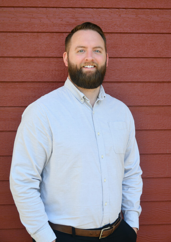 Smiling bearded man in a light button-down shirt standing against a red wooden wall