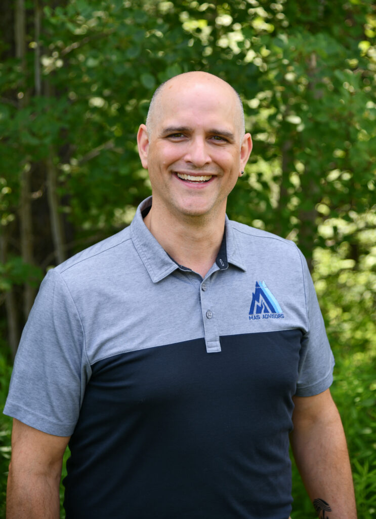 Smiling bald man in a gray and navy MAS Advisors polo shirt outdoors among trees
