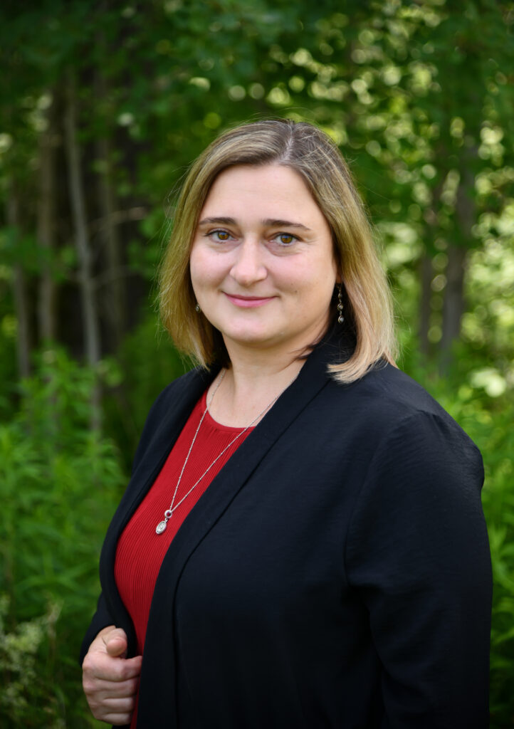 Smiling woman with shoulder-length blonde hair wearing a black blazer and red top, standing in a wooded area