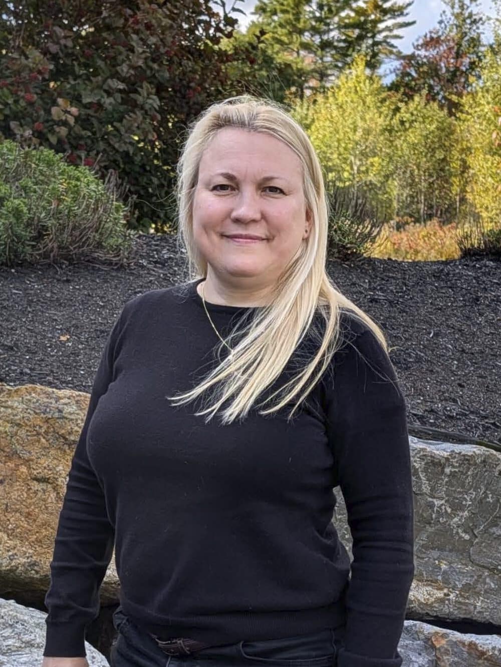Blonde woman in black clothing standing in front of large rocks and autumn foliage