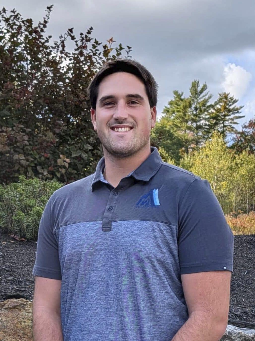 Smiling man in a blue polo shirt standing outdoors in front of autumn foliage and rocks