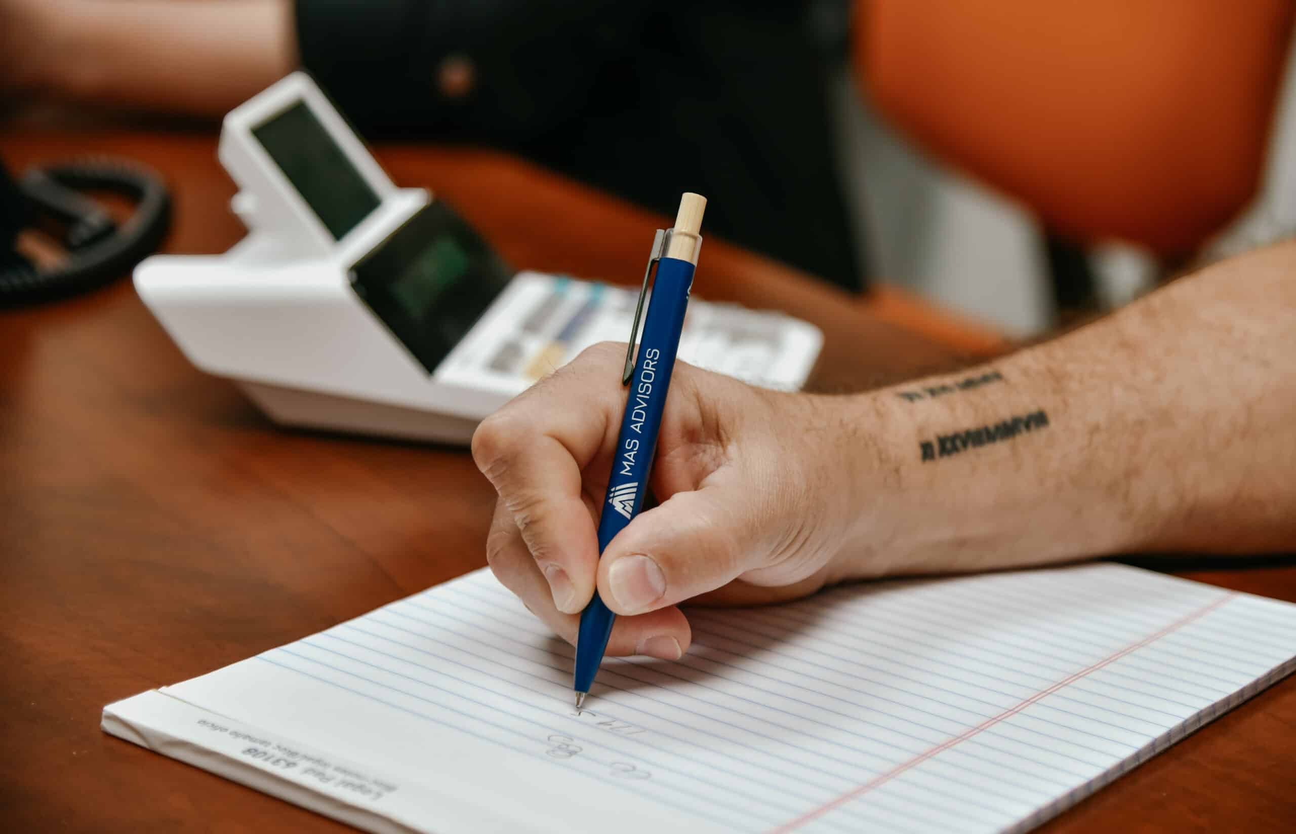 Hand writing in a lined notebook with a blue pen beside a calculator